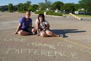 Two ladies sit on cement in front of sidewalk chalk